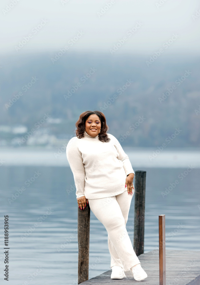 African female adult relaxing by misty lake in cozy sweater outfit on overcast day African female adult relaxing by misty lake in cozy sweater outfit on overcast day