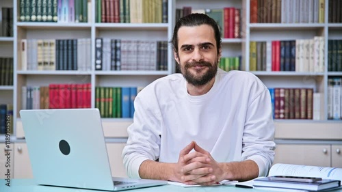 Young handsome bearded man typing on laptop, looking at camera, sitting in office library