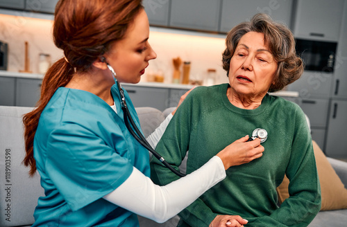 A nurse in a blue suit is checking an elderly woman with a stethoscope while she is sitting on the couch at home. The elderly woman is taking a breath to check her lungs.