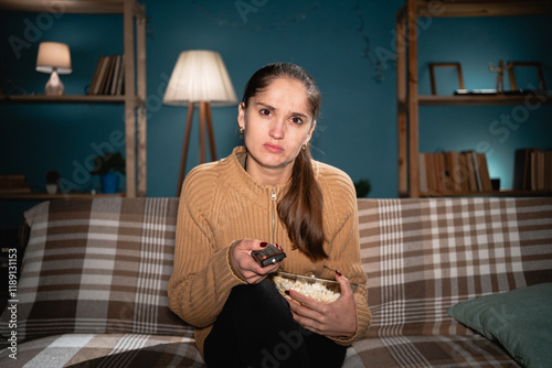 Focused woman holding a remote and a bowl of popcorn, immersed in TV at night. Her intense gaze reflects critical evaluation or curiosity as she engages with the screen content. photo