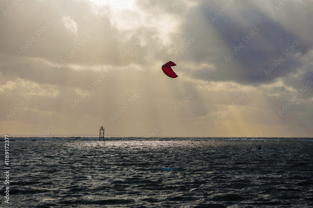 A kitesurfer riding the sea using the power of the wind, with a ship and a sky with clouds behind him.
A kitesurfer riding the sea using the power of the wind, with a ship and a sky with clouds behind him.