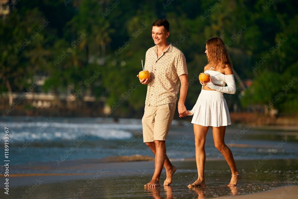Fototapeta premium Romantic couple stroll barefoot on sandy beach, man and woman in summer clothes hold hands enjoying sunset. Pair with tropical drinks walk by sea, honeymoon vacation mood on exotic island getaway.