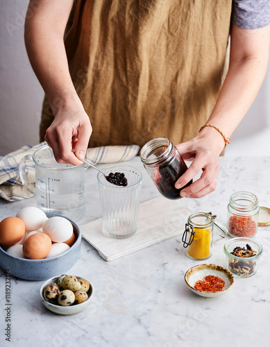 A person prepares ingredients on a marble countertop, measuring out dried berries into a glass while surrounded by eggs, small jars of spices, and baking supplies photo