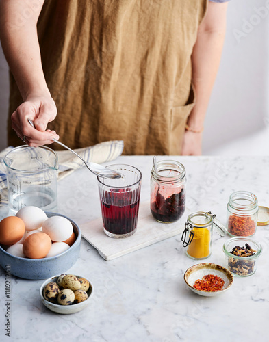 A person in an apron delicately stirs a glass of purple liquid while surrounded by jars of colorful ingredients, eggs, and bowls on a marble countertop photo