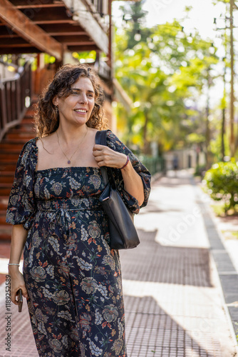Confident woman walking down a sunny street with a handbag and a smile