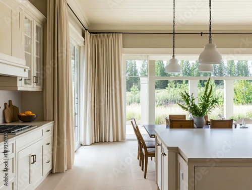 Bright kitchen with creamy cabinets and long beige curtains. Large windows look out onto a green landscape. Three pendant lights hang above a kitchen island.