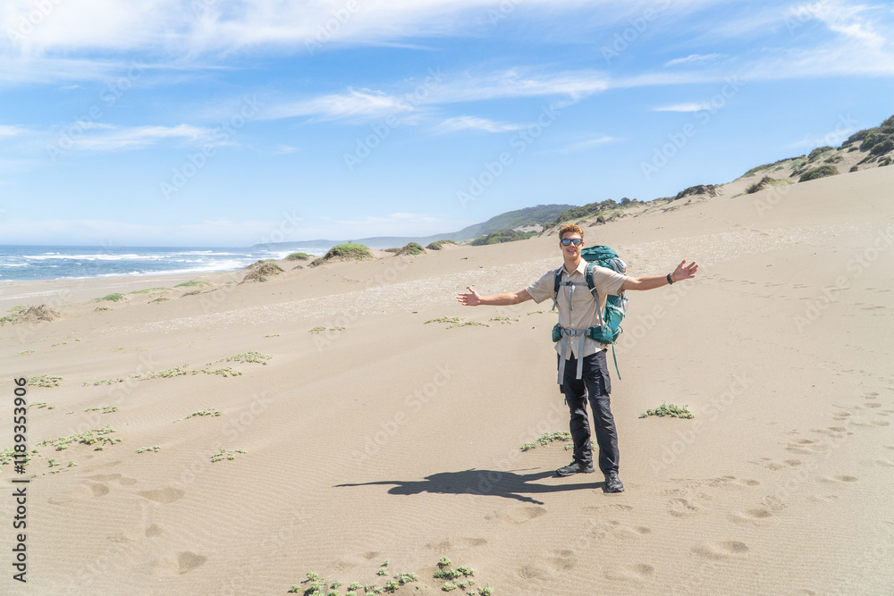 A young backpacker posing for a photo on a sandy beach, wearing a loaded backpack and enjoying the coastal scenery under a clear sky.