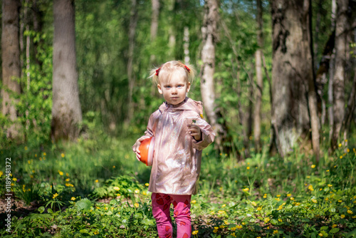 Obraz na płótnie A little girl of 2-3 years old stays in the forest and enjoys nature. Background of a green forest. The girl holds tightly in her hand an orange jar in which she collects the flowers she likes. w salonie