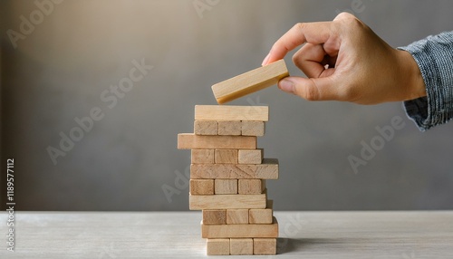 A hand lifts the last block as a precariously stacked wooden block tower is in danger of collapsing.
