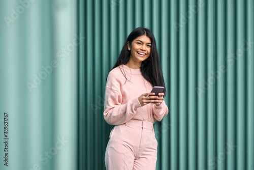 Happy Asian teen girl wearing pink outfit using mobile phone apps outdoors looking at camera. Positive smiling pretty female model holding smartphone laughing standing at blue wall background.