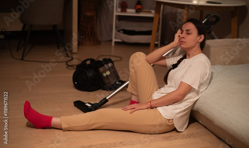 A woman in casual clothing rests while sitting on the floor, looking tired after vacuuming. The room is cozy, with modern decor, and a vacuum cleaner is nearby. The setting is relaxed and homely. photo