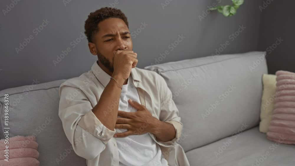 Young man coughing while sitting on a sofa in a modern living room with a serious expression Young man coughing while sitting on a sofa in a modern living room with a serious expression