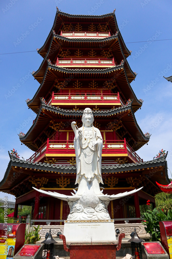 A detailed close-up of a magnificent white Guan Yin statue, positioned in front of a vibrant red and gold Chinese pagoda. A detailed close-up of a magnificent white Guan Yin statue, positioned in front of a vibrant red and gold Chinese pagoda.