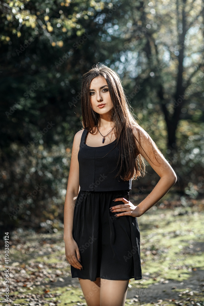young woman with long dark hair standing on the street in sunny weather. She wears a black sleeveless dress and wears a necklace with a pendant. young woman with long dark hair standing on the street in sunny weather. She wears a black sleeveless dress and wears a necklace with a pendant.