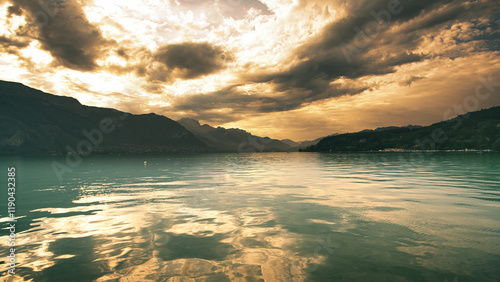 A serene lake reflecting golden hues of a dramatic cloudy sky, surrounded by mountains and lush hills, creating a peaceful and atmospheric natural landscape.
