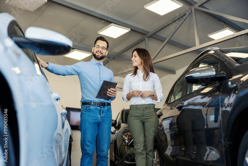 Car seller with tablet in hands showing luxury car for a female customer at car salon.