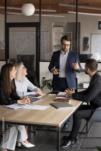 Businessman standing and addressing group of professionals seated at conference table, giving instructions or guiding formal discussion with shareholders, planning, making commercial offer for client