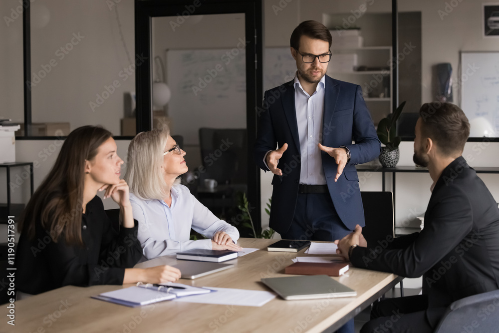 Businessman dressed in suit explaining or presenting information, standing in front of group of business partners gathered in conference room for planning collaboration, partnership or conclude deal Businessman dressed in suit explaining or presenting information, standing in front of group of business partners gathered in conference room for planning collaboration, partnership or conclude deal
