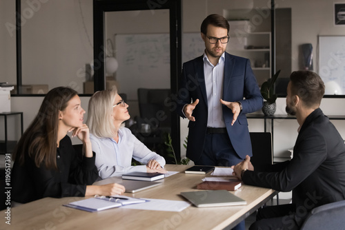 Businessman dressed in suit explaining or presenting information, standing in front of group of business partners gathered in conference room for planning collaboration, partnership or conclude deal