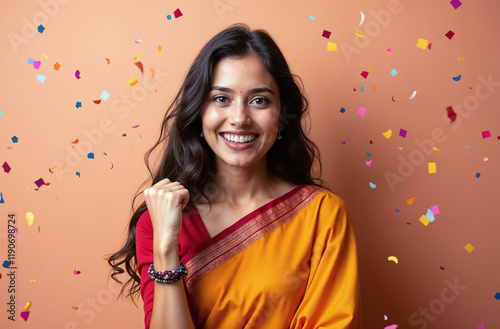 Happy successful young indian woman in a saree, celebrating with her fist raised, isolated on a solid background with confetti