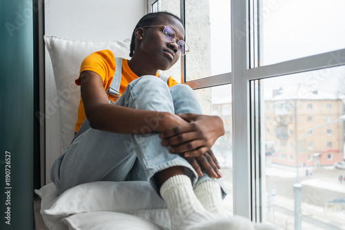 Obraz na plátně Young black woman sits pensively by her window on a winter day, her gaze lost in
