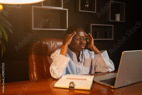 Young businesswoman having a bad headache while working on a laptop late at night, touching temples, suffering from migrain because of stress and overwork, sitting at desk in dark office photo