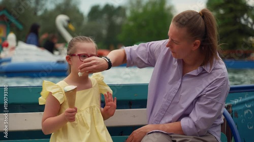 Child with Down syndrome gestures expressively while holding ice cream, as her mother gently wipes her mouth with napkin. They sit by water and colorful attractions. Concept care and inclusion photo