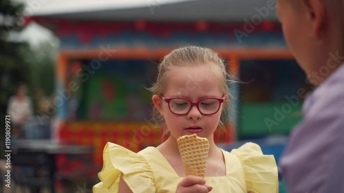 Little girl with Down syndrome enjoys ice cream while talking to her mother in amusement park. Her face is focused on conversation, with colorful attractions in blurred background. Concept inclusion photo