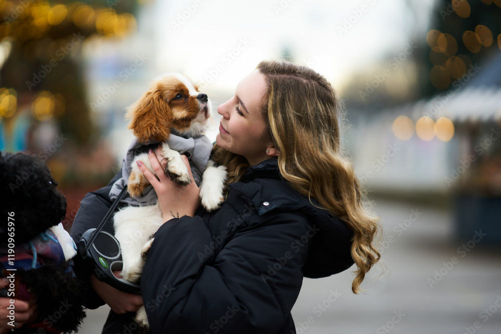 Enjoying a chilly day outdoors, a woman holds a playful puppy while surrounded by holiday decorations and festive lights Enjoying a chilly day outdoors, a woman holds a playful puppy while surrounded by holiday decorations and festive lights