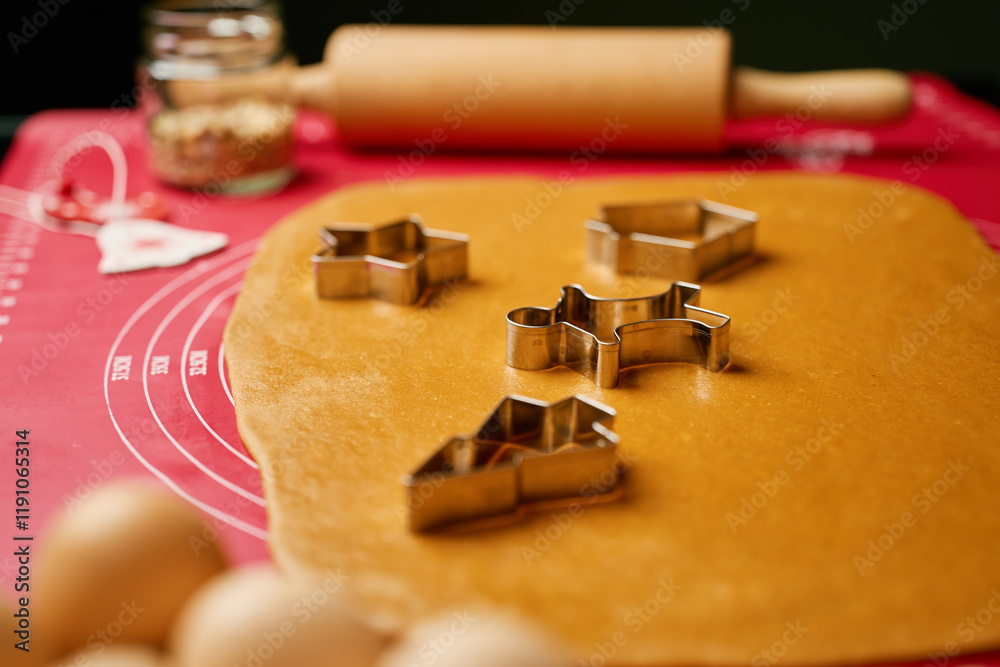 Rolled-out gingerbread dough with metal cookie cutters in shape of stars, houses, and gingerbread men on red mat Rolled-out gingerbread dough with metal cookie cutters in shape of stars, houses, and gingerbread men on red mat