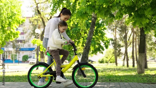 Woman and a child are riding a yellow bike in a park. The woman is holding the child's hand and guiding him. Scene is happy and playful. happy family. girl mother park. woman child kid walk happy