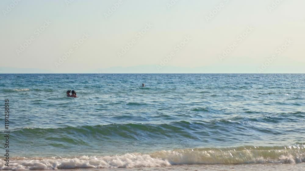 People swimming in clear Aegean Sea with distant mountains visible through haze on horizon under soft summer sunlight.