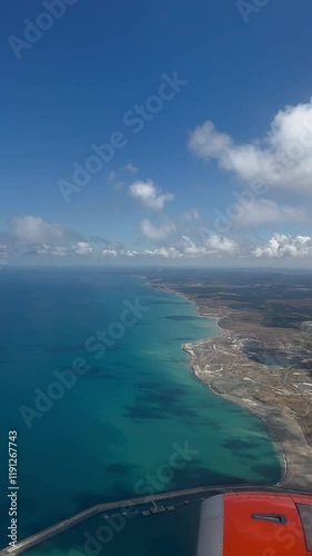 Aerial view of a coastline with turquoise waters, white clouds, and rugged landscapes, taken from an airplane. The image captures the contrast between the vibrant sea and the arid land