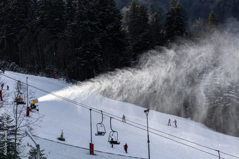 Winter ski resort. Beautiful view with clouds of snow created by the snow cannon machine. Tourist skiing on slopes. Winter sports.