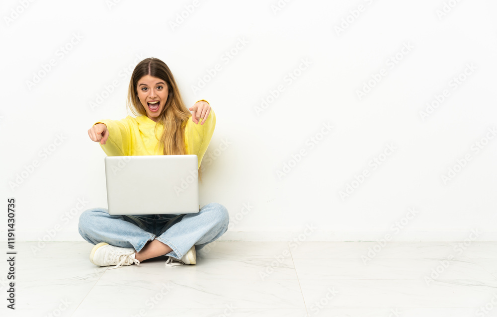 Young woman with a laptop sitting on the floor surprised and pointing front Young woman with a laptop sitting on the floor surprised and pointing front
