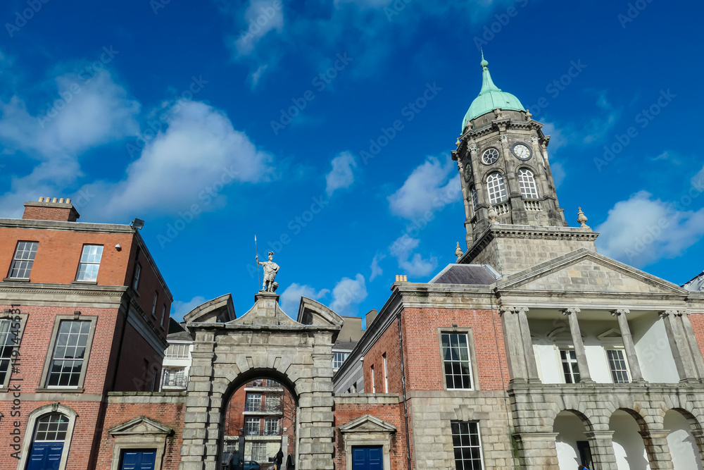 Obraz premium Dublin Castle's Bedford Tower in Ireland. Green dome and clock faces, stands prominently against blue sky. Stone gateway topped by statue of Justice leads into castle grounds. Georgian brick buildings