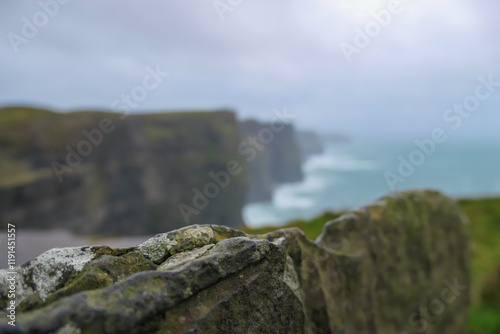 Selective focus on rock formation in dramatic coastal scene of Cliffs of Moher in Ireland. Steep cliff drops directly into Atlantic Ocean. Misty and moody atmosphere. Blurred background of landscape photo