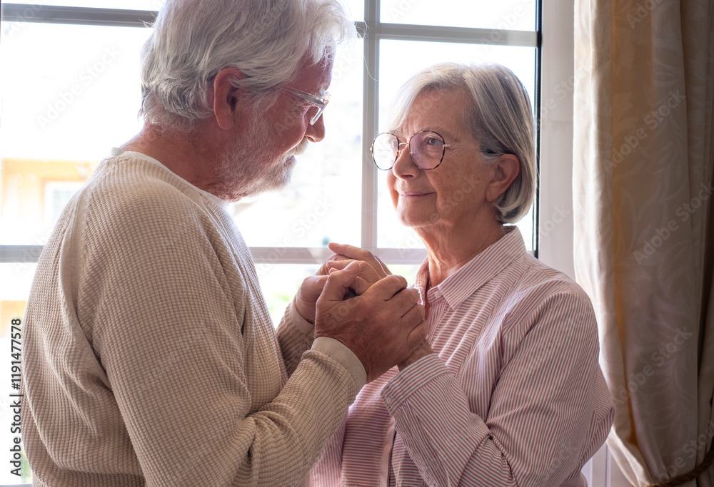 Obraz premium Senior Caucasian couple holding hands in front of the window, an elderly man and woman looking into each other's eyes supporting and helping