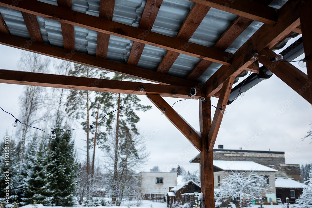 Fototapeta premium A wooden terrace with a corrugated metal roof and string lights, set against snow covered trees, buildings, and a prominent rectangular structure.