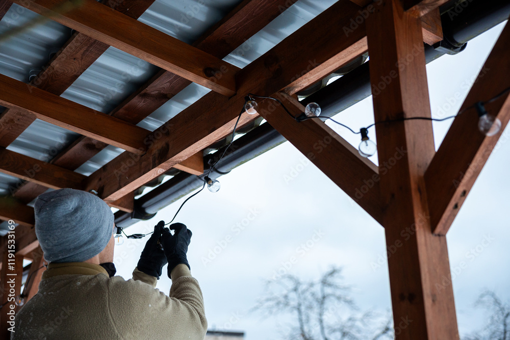A person in a gray beanie and gloves installs LED bulbs on a wooden terrace structure with a corrugated metal roof, set against bare trees and a cloudy sky. A person in a gray beanie and gloves installs LED bulbs on a wooden terrace structure with a corrugated metal roof, set against bare trees and a cloudy sky.