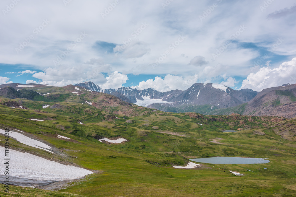 Fototapeta premium Scenic awesome landscape with big snowfield and alpine lake among green hills and rocks with view to glacier and large snow-capped mountain top far away under clouds in blue sky in changeable weather.