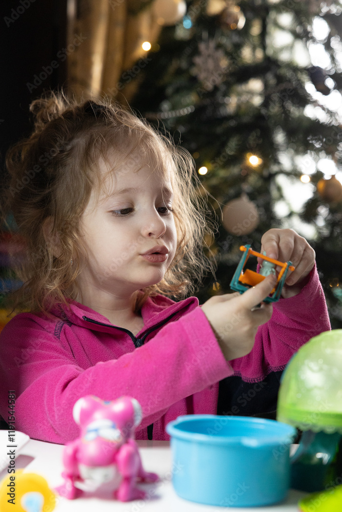 Fototapeta premium The child plays with toys. A little girl sits at a table, against the backdrop of a Christmas tree. Toys are located on the table.