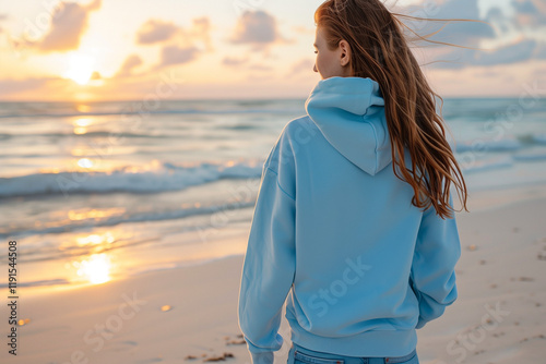Sky Blue Hoodie Mockup: Stylish Female Walking Along Sandy Beach with Ocean Waves in Background, Casual Fashion Concept for Apparel Design photo