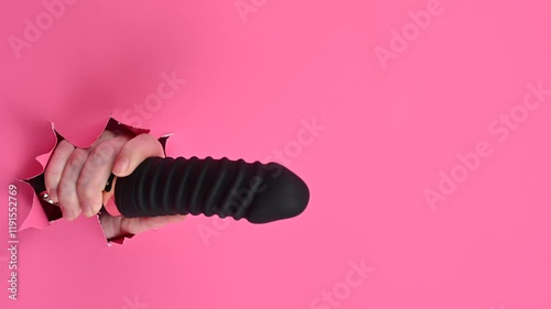 Female hand sticking out of pink paper background with black textured vibrator. Copy space. 