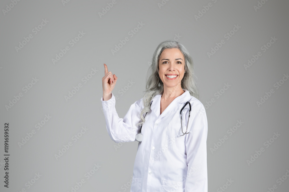 A middle aged woman wearing a white coat stands confidently, pointing upwards with a smile, copy space