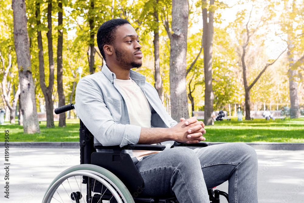 Fototapeta premium Full length of peaceful black handicapped guy in wheelchair having walk at urban park in autumn. Paraplegic African American man deep in thought, spending time outdoors in fall