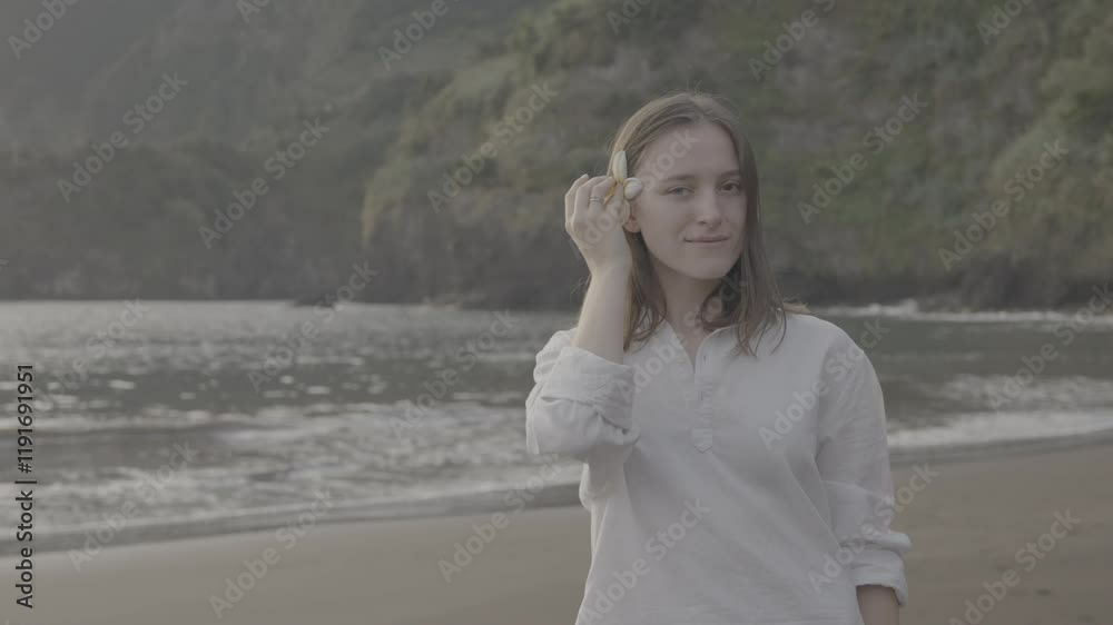 Young woman holding tropical flower near her face while smiling on beach on Madeira, Portugal, at sunrise (SLOG3) Young woman holding tropical flower near her face while smiling on beach on Madeira, Portugal, at sunrise (SLOG3)
