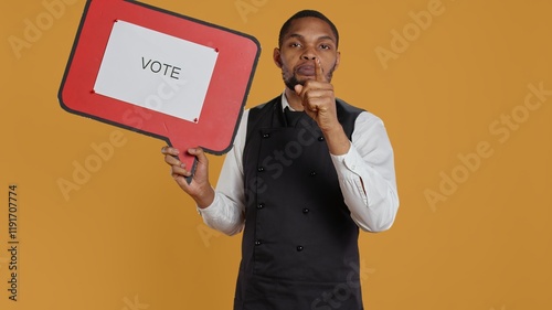 Catering employee showing a message to vote on red speech bubble, presenting the cardboard icon to do a commercial for politics. Waiter in uniform works on customer service. Camera B. photo