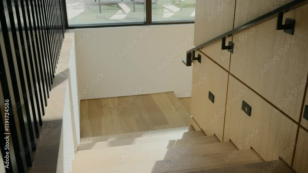 Modern staircase with wooden steps and sleek, black handrail. Wooden wall on right side of staircase and natural light streaming through large window at top of stairs