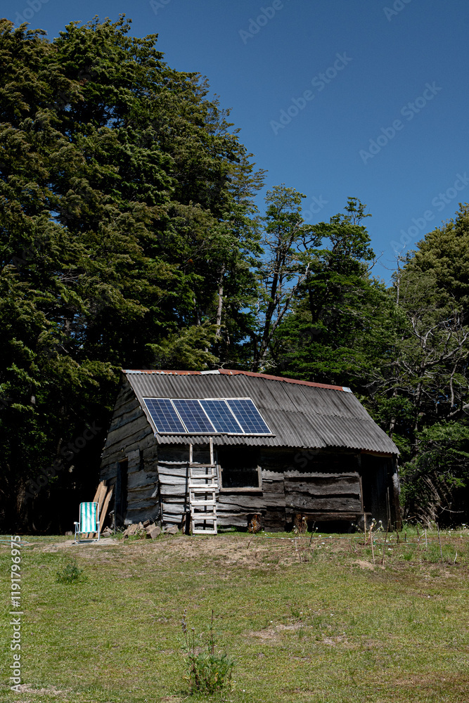 Fototapeta premium A rustic wooden cabin with solar panels on its roof, nestled in a clearing surrounded by lush green trees under a bright blue sky.